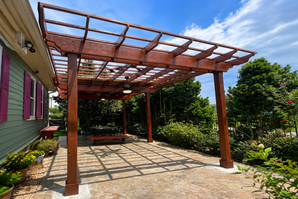 Custom stained pine pergola patio cover installed over a backyard stone patio in South Louisiana with decorative rafters and natural sunlight.