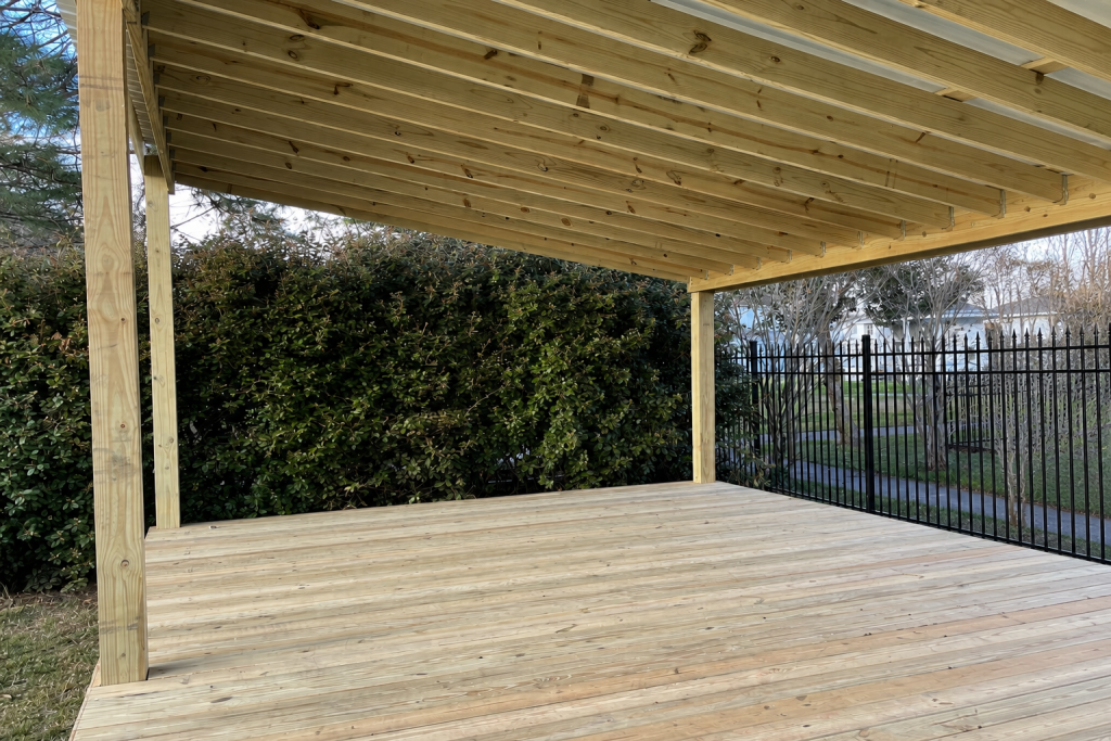 Newly completed pressure-treated wood covered deck with exposed shed roof framing, black aluminum fence, and hedge backdrop in Covington Louisiana.