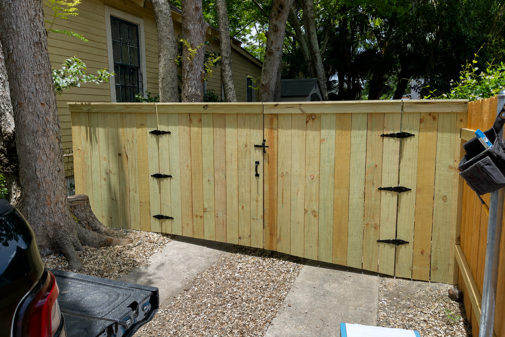 Pressure-treated pine double drive gate with black decorative hinges and latch installed between privacy fence sections in a shaded residential backyard.